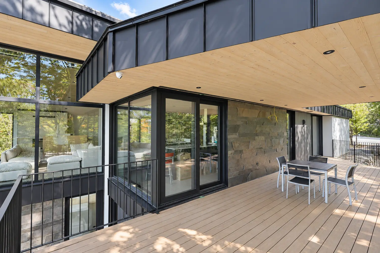 Covered deck with a black-framed sliding glass door between a stone wall and the glazed living room, with an outdoor table and chairs