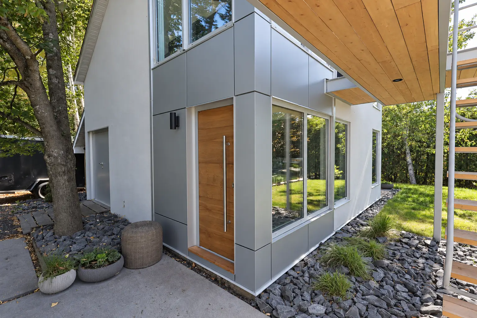 Wood-finish entry door with a long vertical bar handle set in grey metal cladding, next to corner windows under a wood-toned soffit
