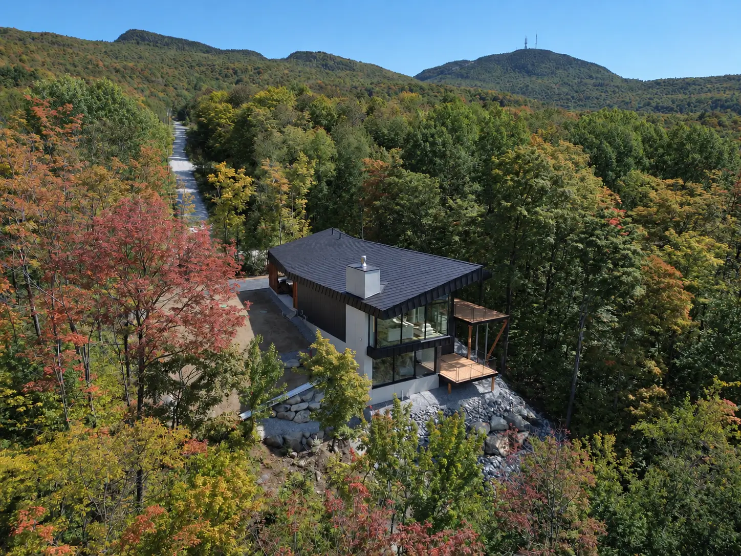 Aerial view of a dark-roofed house with floor-to-ceiling glazing, surrounded by autumn forest with hills beyond