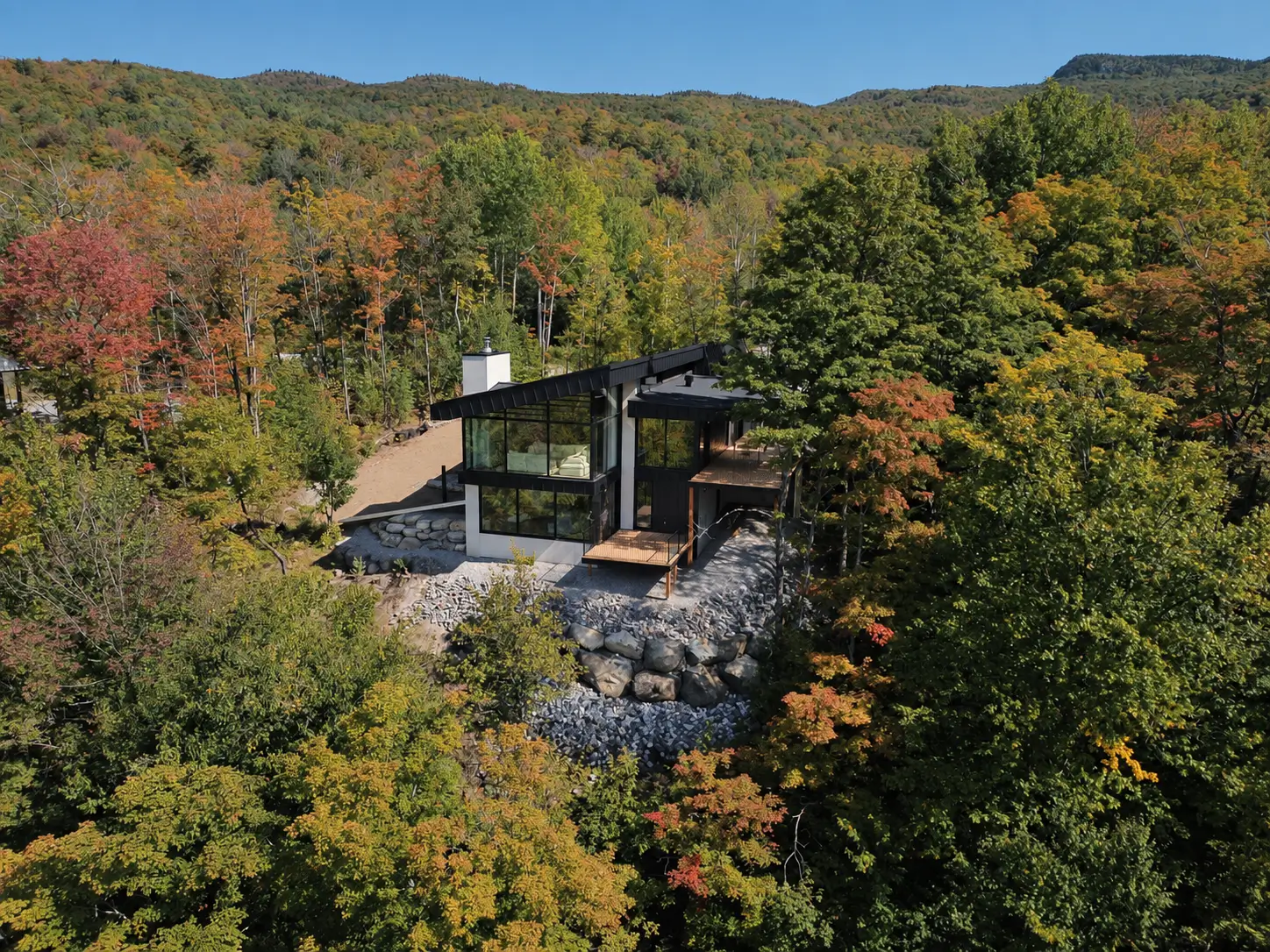 Aerial view of the glazed front of the house and its decks on a rocky slope, surrounded by autumn forest
