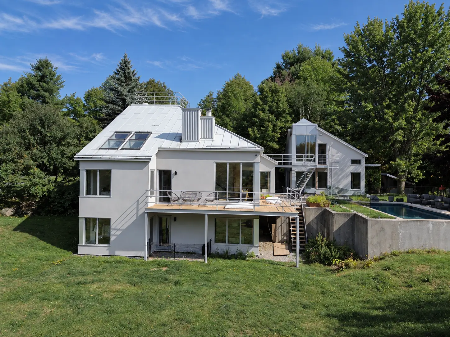 Garden view of a light grey house with a white metal roof and grey-framed windows, with a raised deck, a glazed annex and a swimming pool