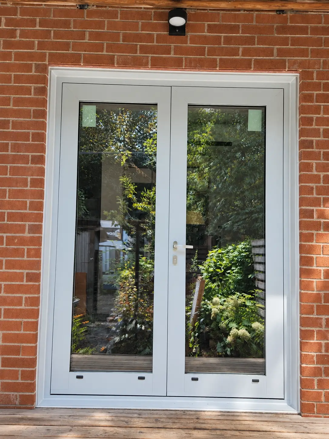 Glazed double door in a red-brick wall opening onto a wood deck, with the garden reflected in the glass