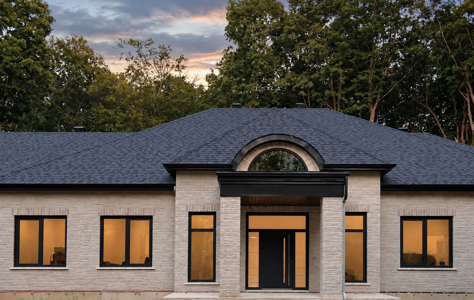 Stone-clad house at dusk with a columned entry portico, a dark entry door with glazed sidelights and dark-framed windows lit from inside