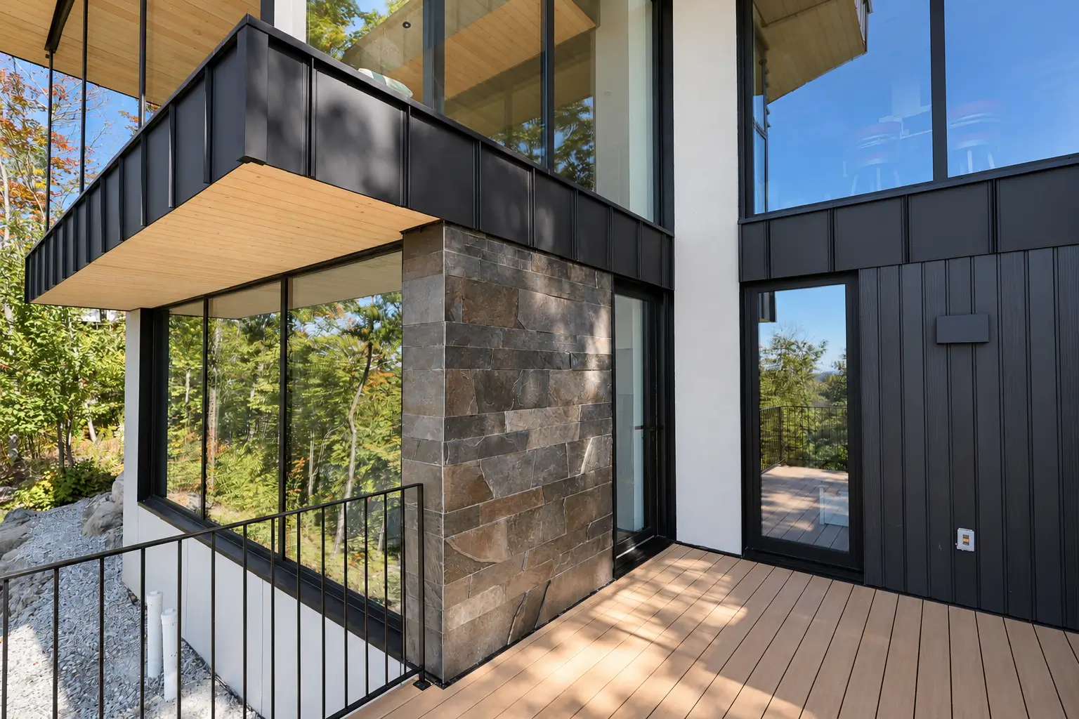 Lower level of the house where black-framed windows meet stone cladding, a white wall and dark metal siding beside a deck