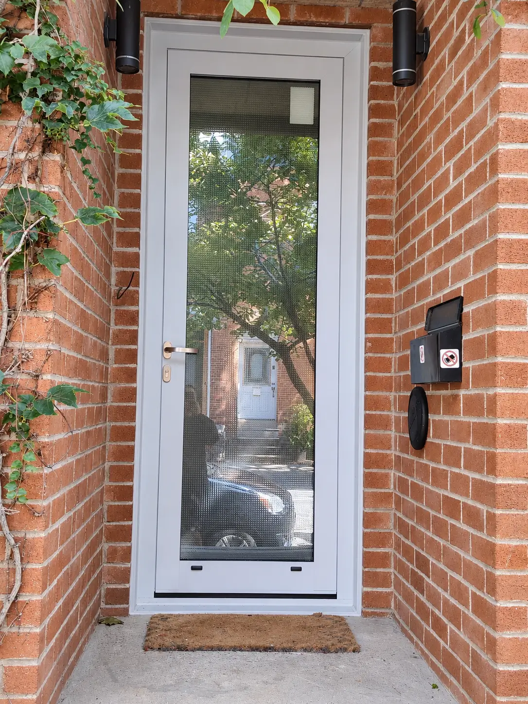 Entry door with full-height textured glass in a red-brick recess, with two black wall lights and a doormat