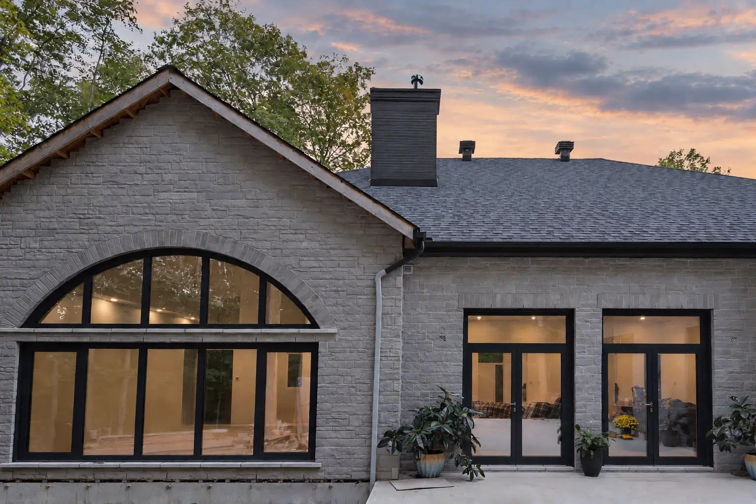 Stone-clad gable at dusk with the arched window wall lit from inside, beside two dark-framed glazed double doors with transoms and potted plants in front