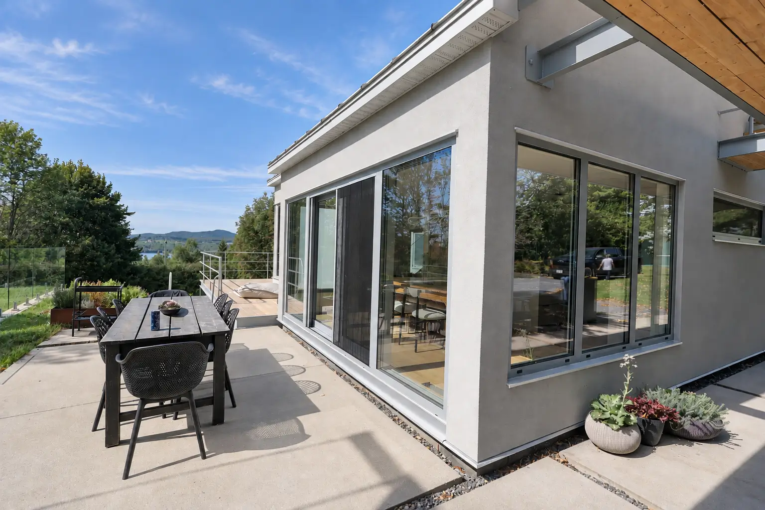 Corner of the house with a large grey-framed sliding glass door and a three-part window beside a terrace dining table, with a lake and hills in the distance